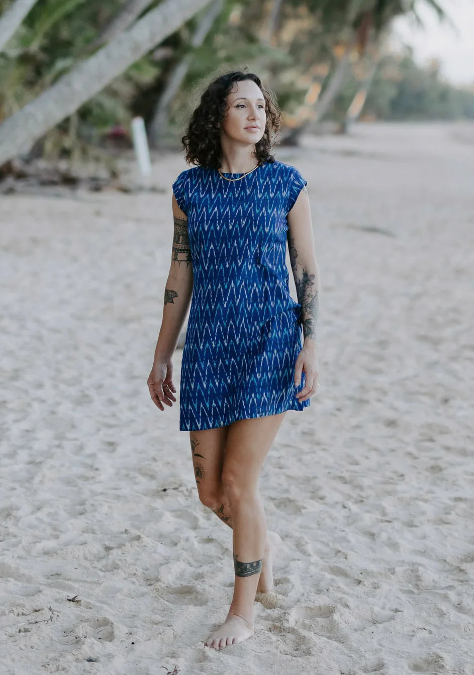 Lady looking out to sea, walking on the beach under palms, wearing a electric blue handloomed cotton shift dress. Above knee, round neck. 