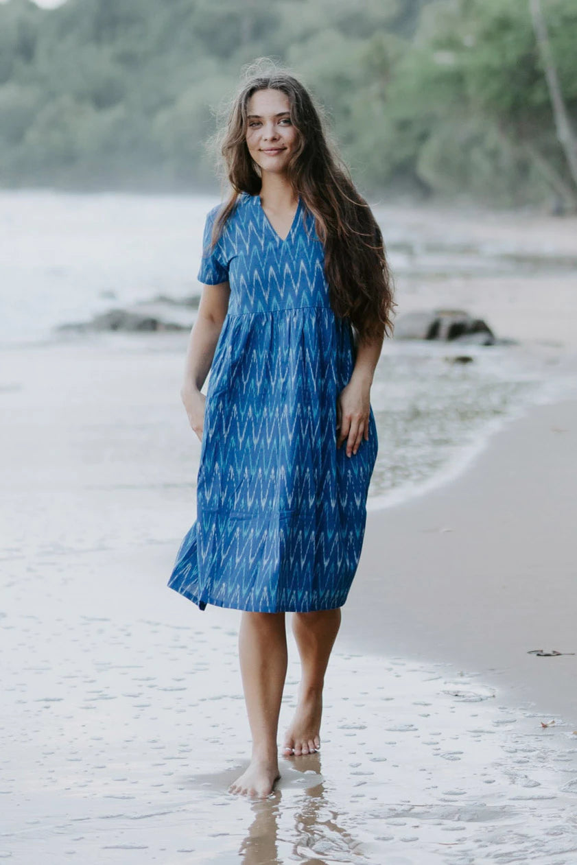 Woman wearing a blue Ikat cotton dress with a V-neckline and gathered waist, walking along the shoreline, showcasing sustainable and ethical fashion