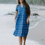 Woman wearing a blue Ikat cotton dress with a V-neckline and gathered waist, walking along the shoreline, showcasing sustainable and ethical fashion