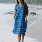 Woman wearing a blue Ikat cotton dress with a V-neckline and gathered waist, walking along the shoreline, showcasing sustainable and ethical fashion