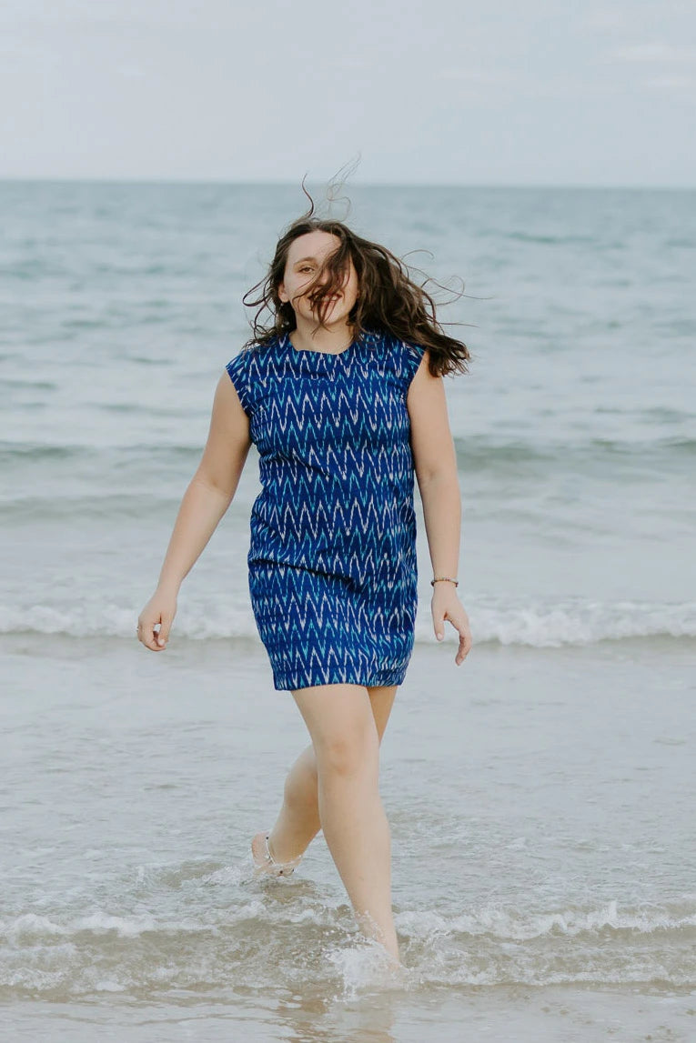 Woman walking in the shallow tide water at the beach, wind in her hair wearing electric blue handloomed cotton dress. above knee, thoughtfully and ethically created at our own studio. Empowering women to support themselves and their families 