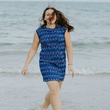 Woman walking in the shallow tide water at the beach, wind in her hair wearing electric blue handloomed cotton dress. above knee, thoughtfully and ethically created at our own studio. Empowering women to support themselves and their families 