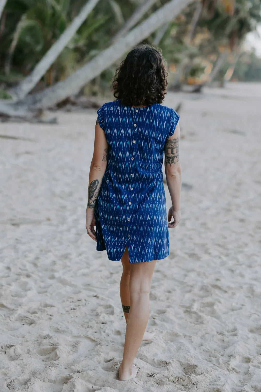 Woman wearing Tara Shirt shift dress walking away under coconut palms in the beach sand. mother of pearl buttons