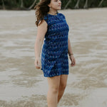 Woman on low tide beach wearing electric blue Tara shirt dress. Wind waving hair. Looking out to sea