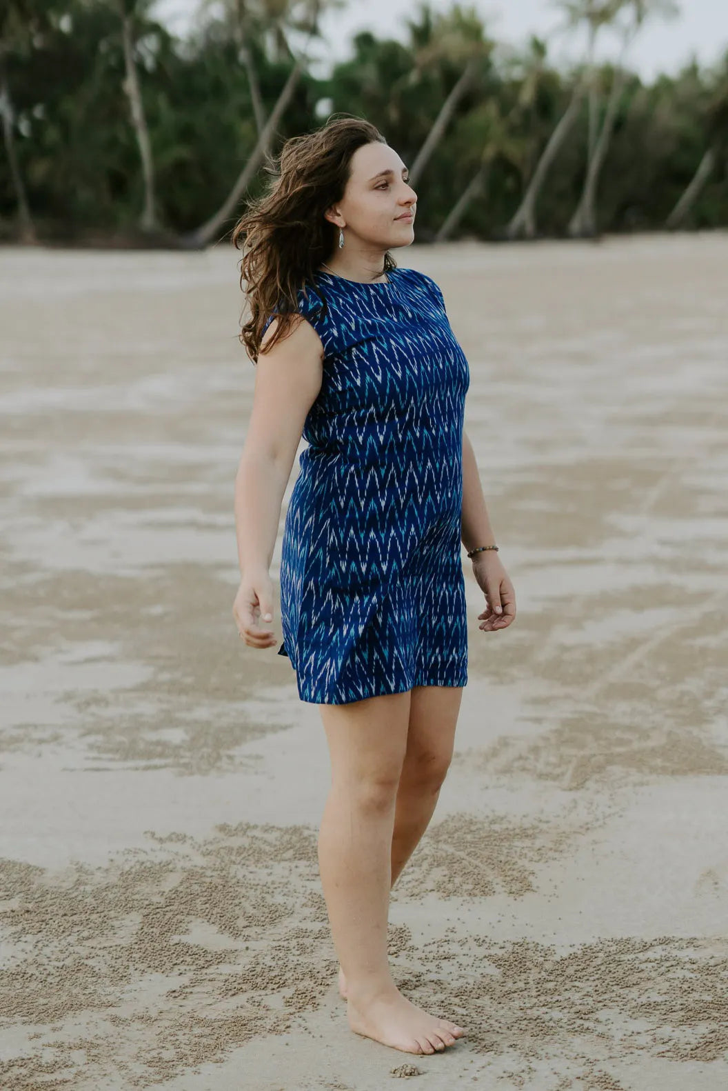 Woman on low tide beach wearing electric blue Tara shirt dress. Wind waving hair. Looking out to sea